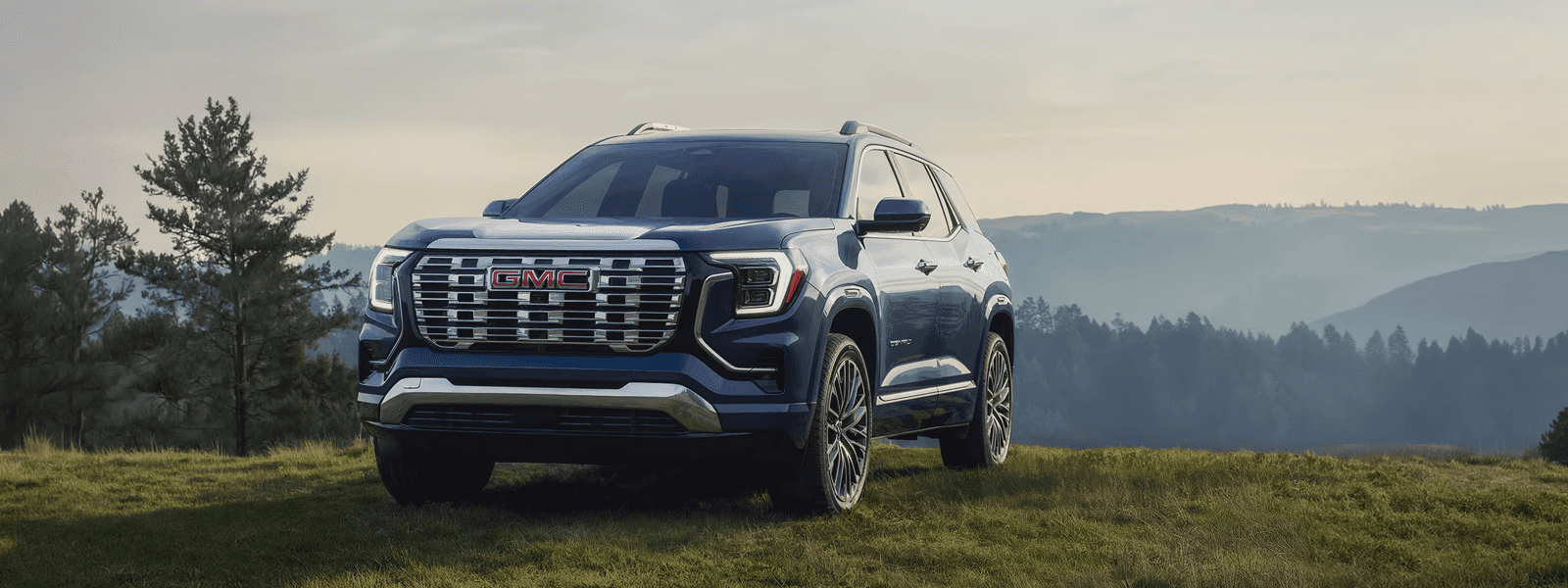 Blue 2026 GMC Terrain Denali driving on a mountain highway showing LED lighting and bold GMC grille design.