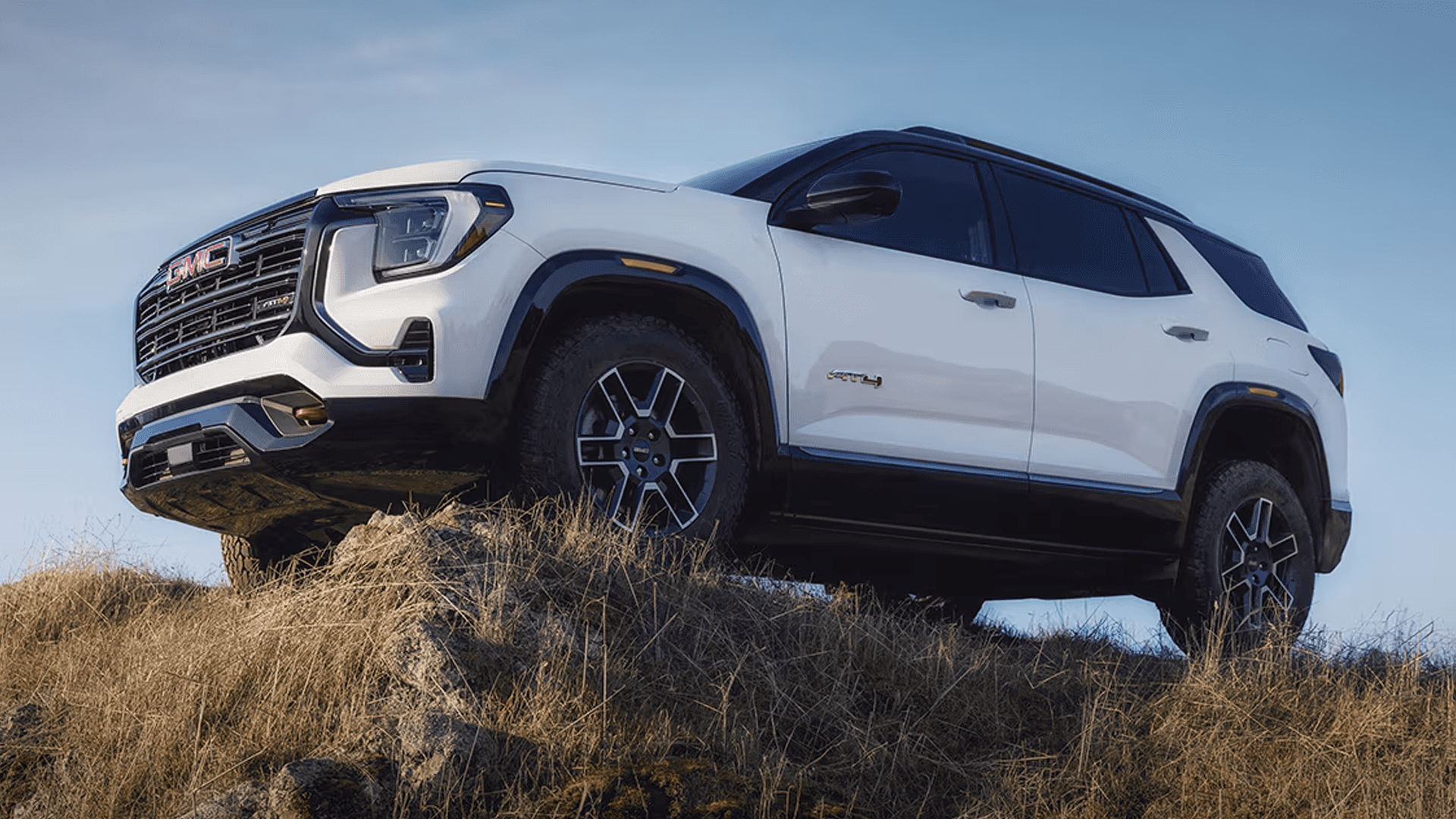 White GMC Terrain AT4 climbing over a grassy ridge with rugged tires and elevated stance in an outdoor setting.