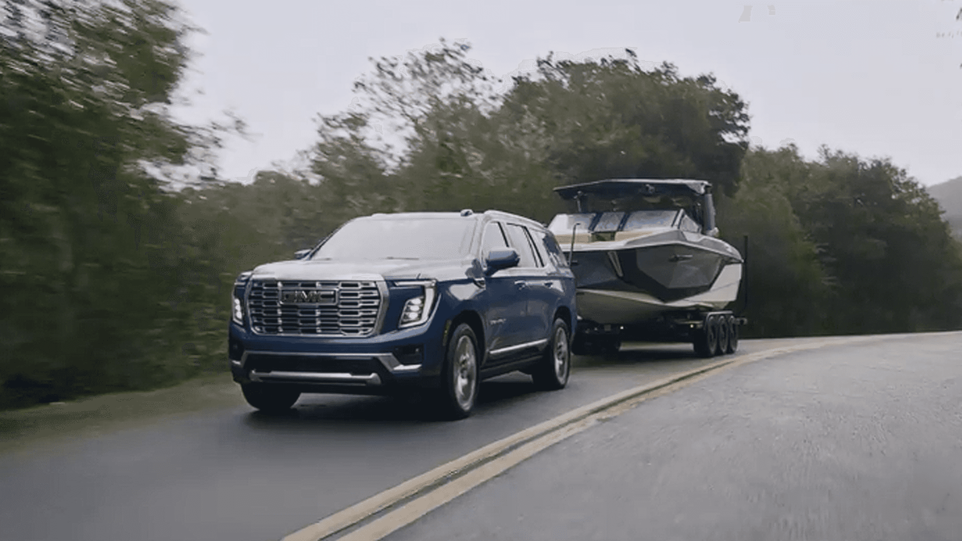 Blue GMC Yukon towing a large boat on a trailer along a wet two-lane road lined with trees.