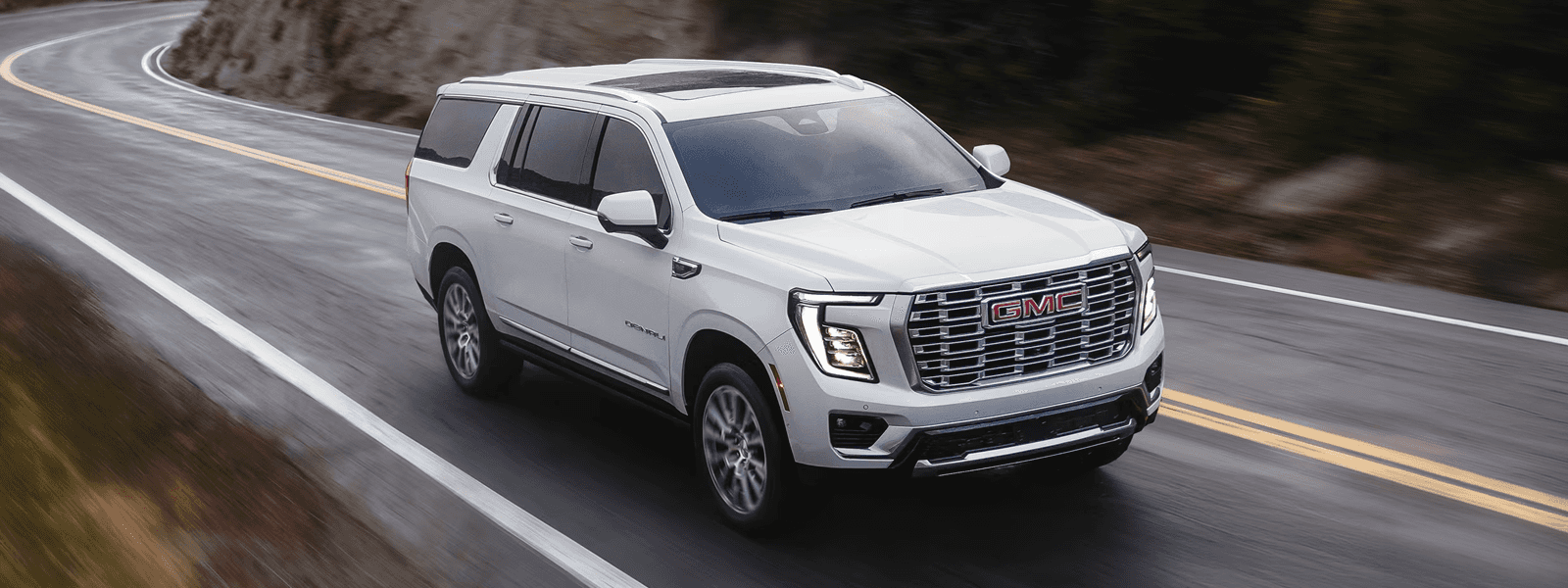 2026 GMC Yukon towing a boat on a mountain road with a lake in the background.