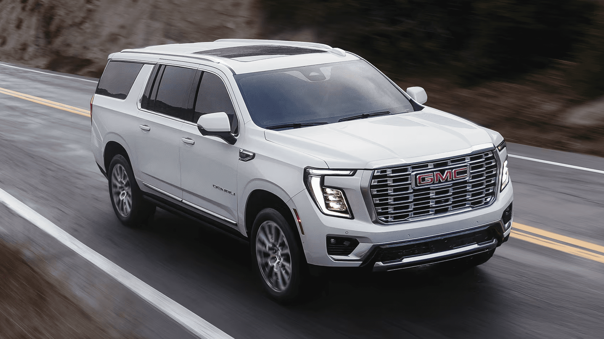 White GMC Yukon driving on a wet road with chrome grille, LED headlights, and roof rails against blurred hillside background