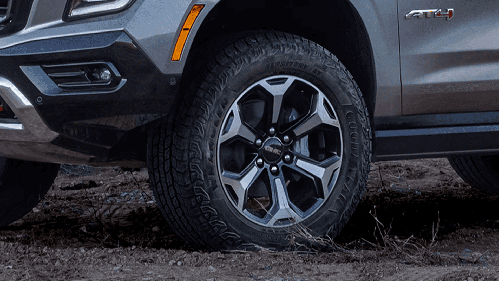 Close-up of SUV front wheel and all-terrain tire on dirt surface with visible tread pattern and alloy wheel design