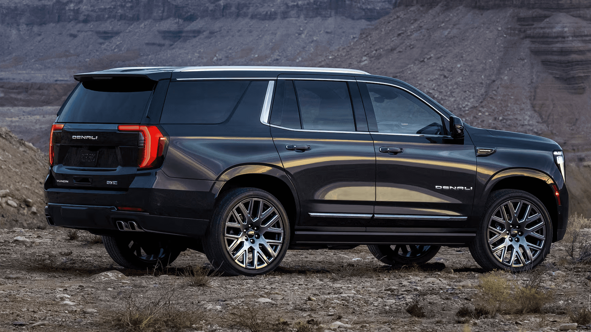 Side view of dark SUV parked on rocky terrain, showing rear LED taillights, large wheels, and Denali badging