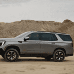 Silver GMC Yukon shown in full side profile driving across sandy terrain with rocky hills and overcast sky in background
