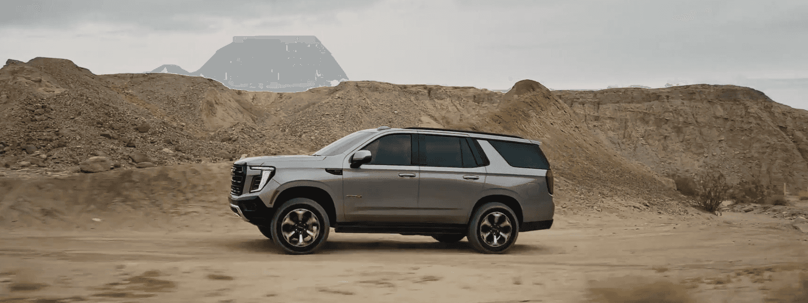 Silver GMC Yukon shown in full side profile driving across sandy terrain with rocky hills and overcast sky in background