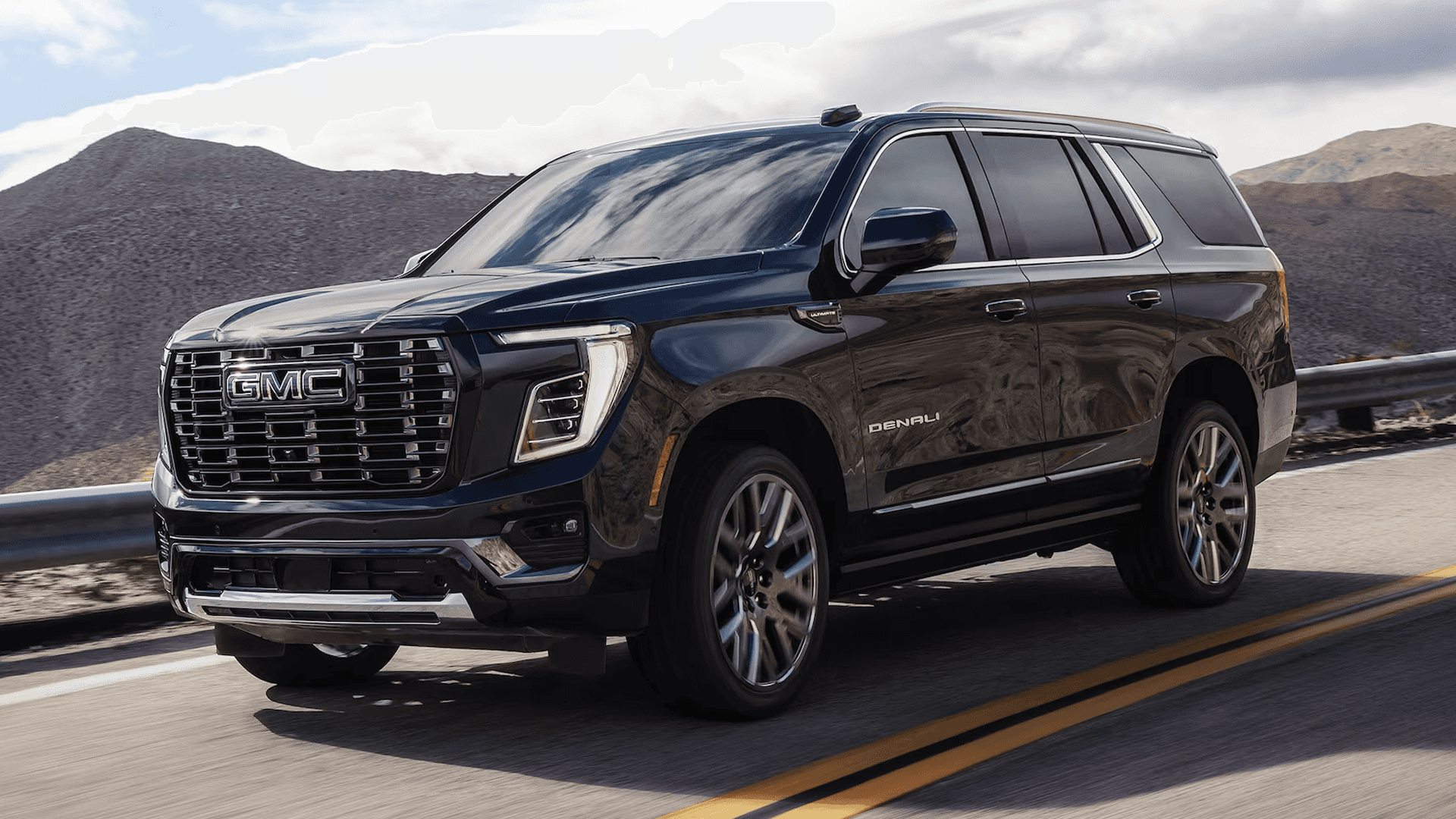 Dark blue GMC Yukon Denali driving on a desert highway with chrome grille, LED lighting, and mountain scenery