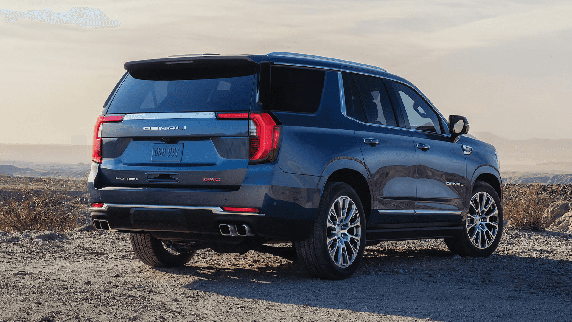 Blue GMC Yukon Denali shown from the rear in desert terrain with LED taillamps, chrome trim, and large wheels