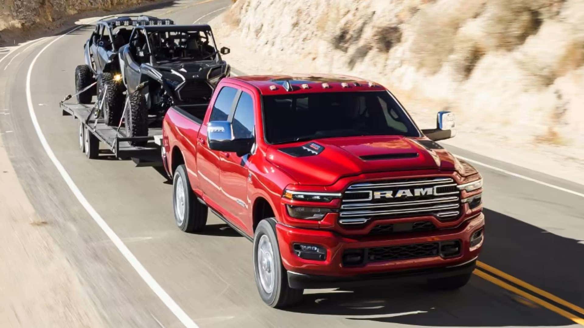 Red 2026 RAM 2500 towing an off road side by side on a trailer along a mountain highway demonstrating heavy duty towing capability