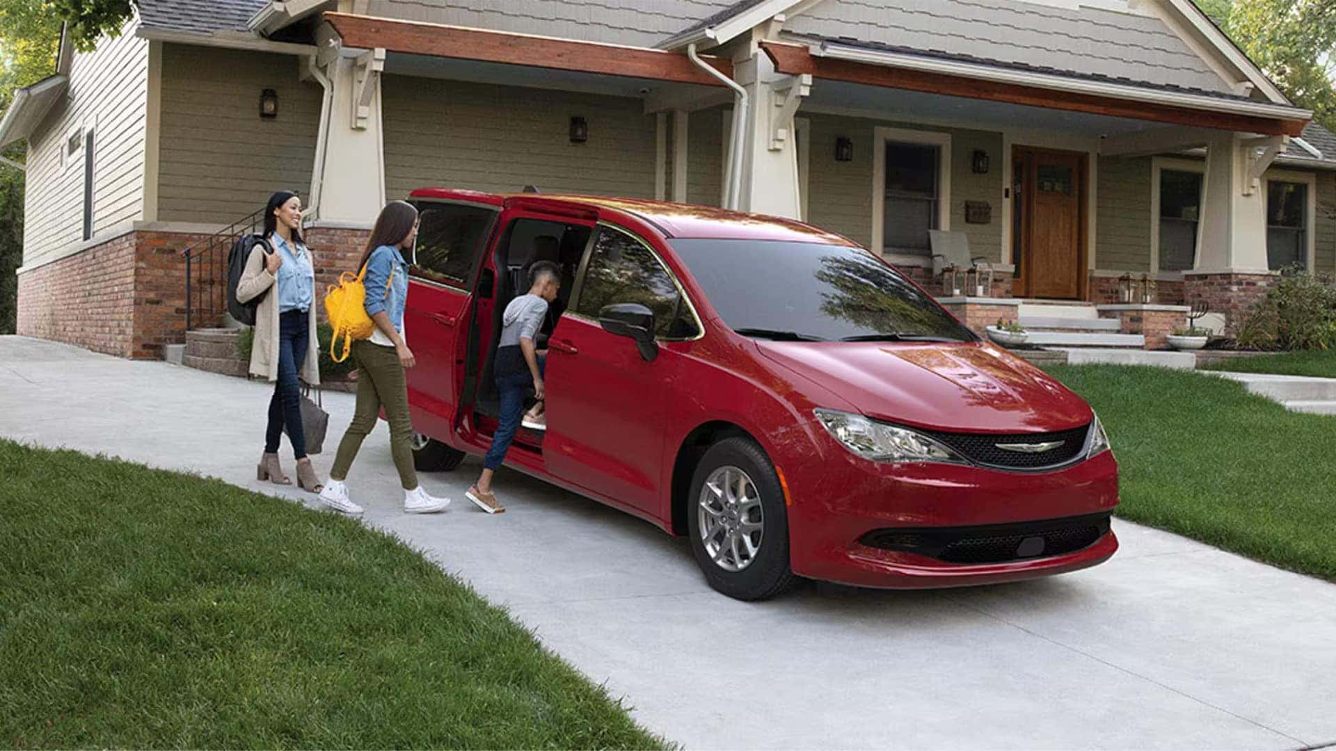 Family entering a red 2026 Chrysler Voyager in a residential driveway, showing power sliding door and exterior design
