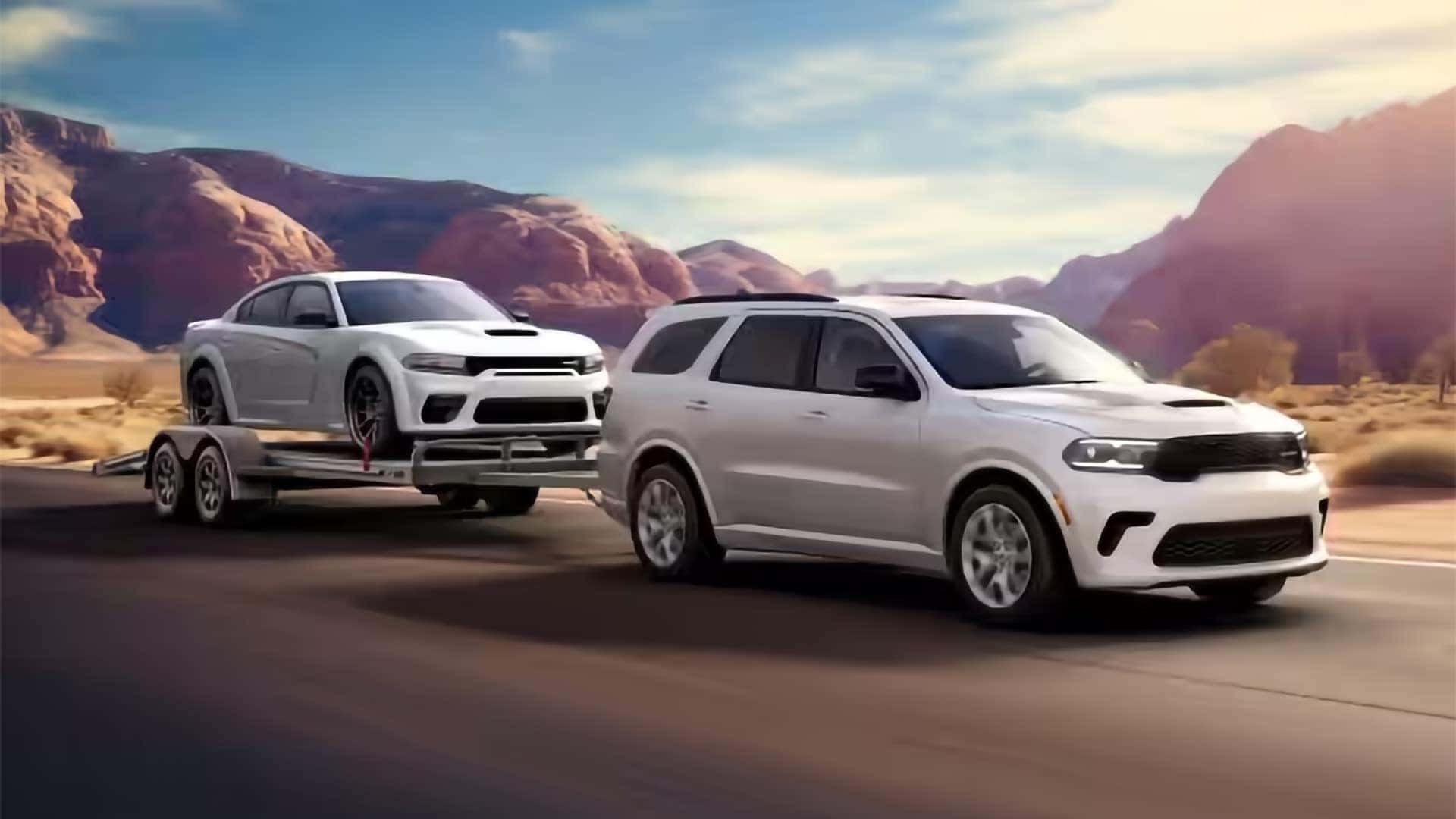 White 2026 Dodge Durango towing a Dodge Charger on a trailer through a desert landscape to show towing strength.