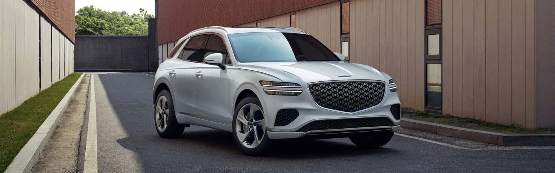 2026 Genesis GV70 luxury SUV in white driving through a modern urban setting, front three quarter view
