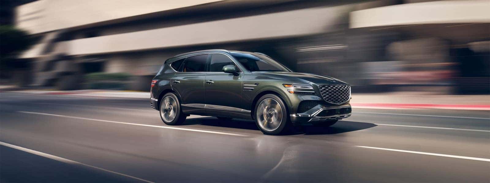 2026 Genesis GV80 luxury SUV driving through a city street in motion with modern architecture in the background