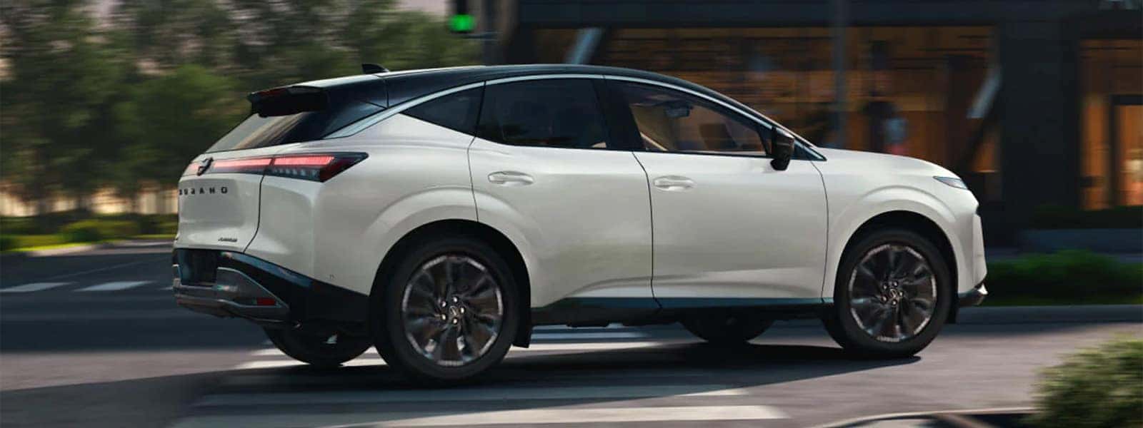 White 2026 Nissan Murano driving through city street, rear three quarter exterior view with black roof and slim LED taillights