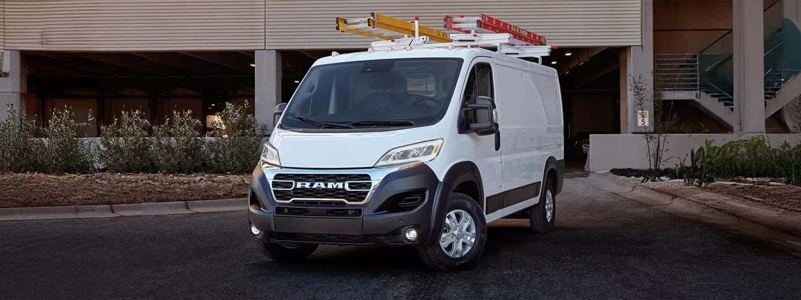 White 2026 RAM ProMaster Cargo Van with roof rack parked outside commercial building, front three quarter view