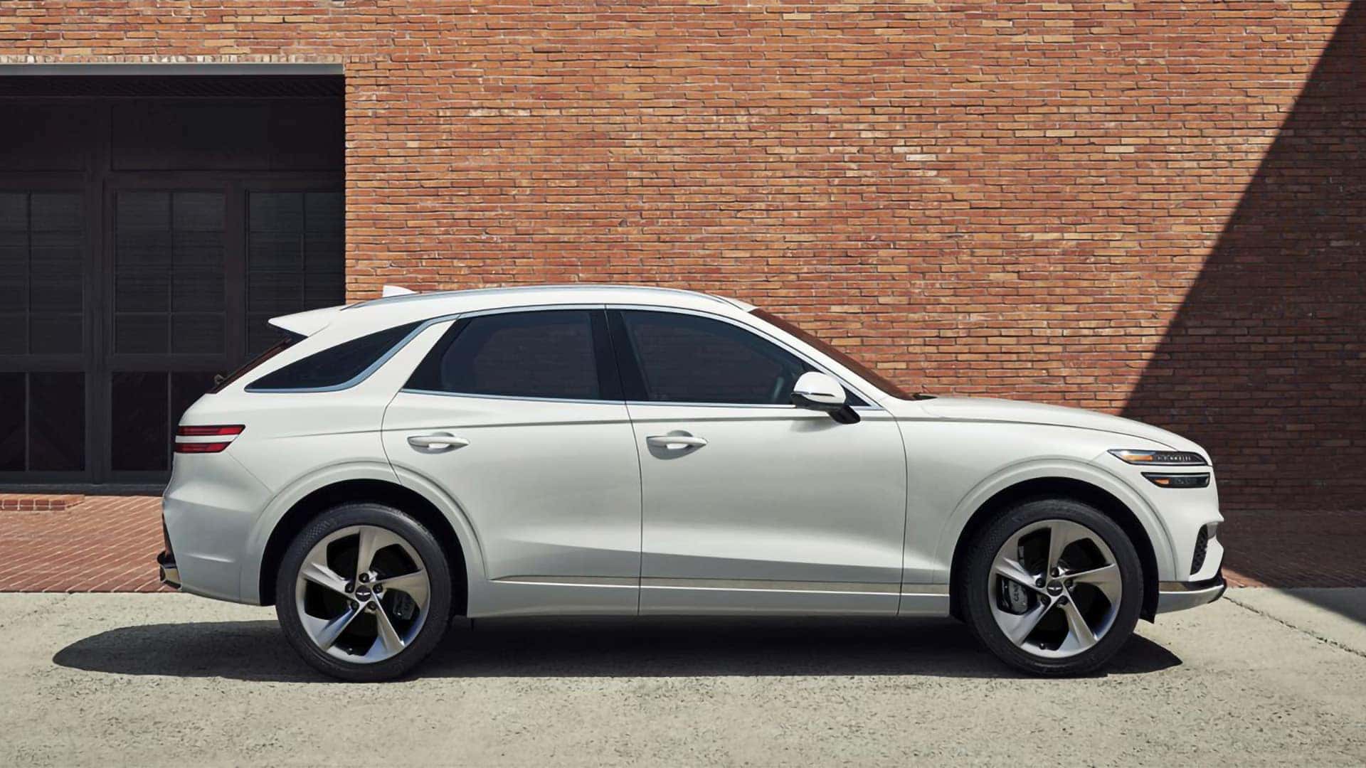 2026 Genesis GV70 side profile parked near a brick wall, showing sleek body lines and alloy wheels