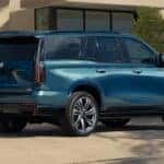 Side rear three quarter view of blue Cadillac Escalade parked in driveway near modern home with large wheels and vertical taillights