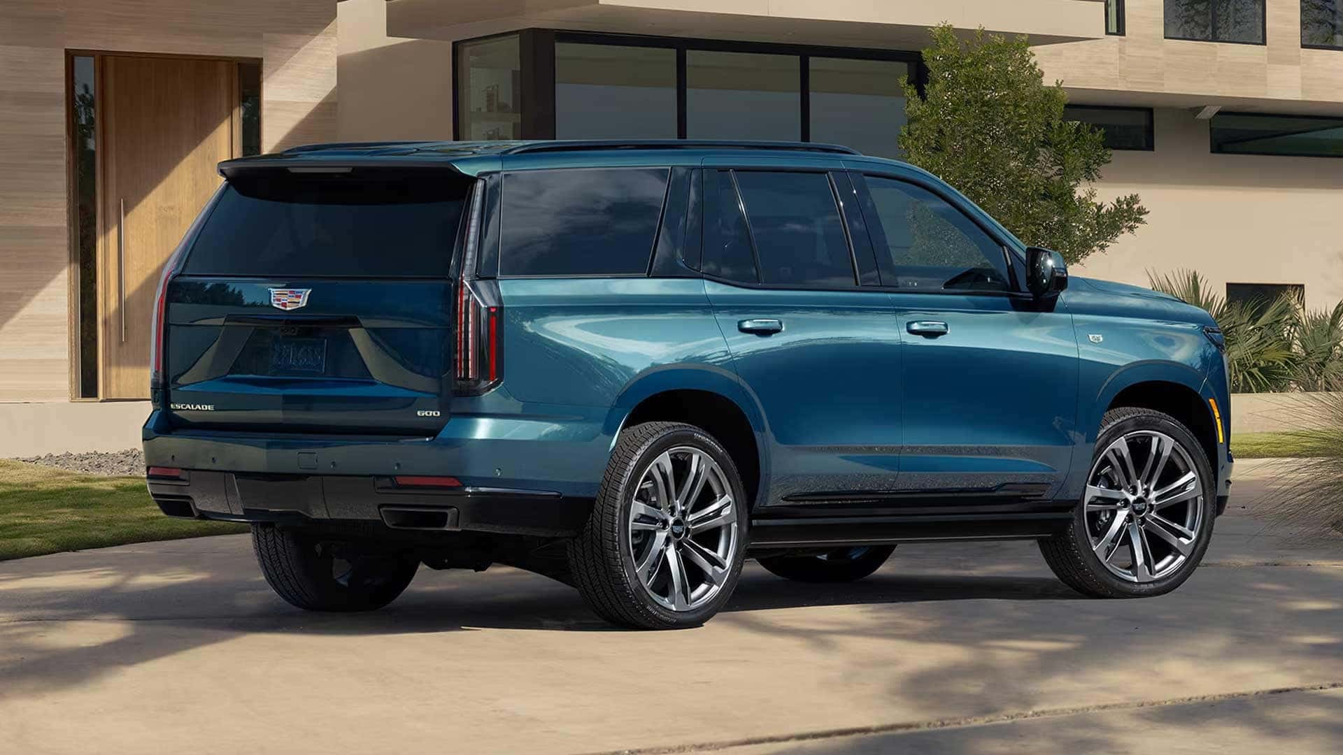 Side rear three quarter view of blue Cadillac Escalade parked in driveway near modern home with large wheels and vertical taillights