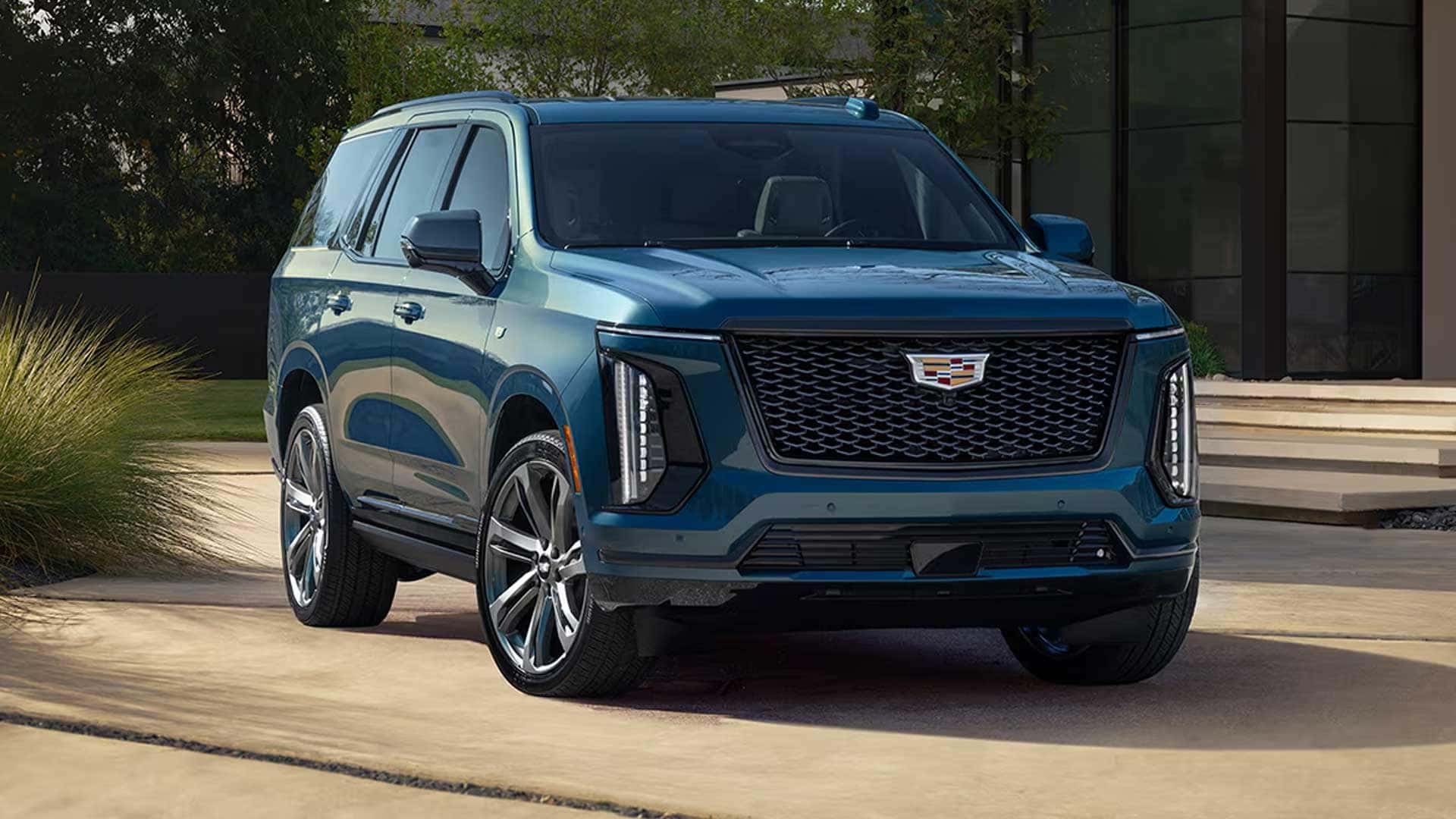 Front three quarter view of blue Cadillac Escalade parked outside modern house showing grille, headlights, and large wheels