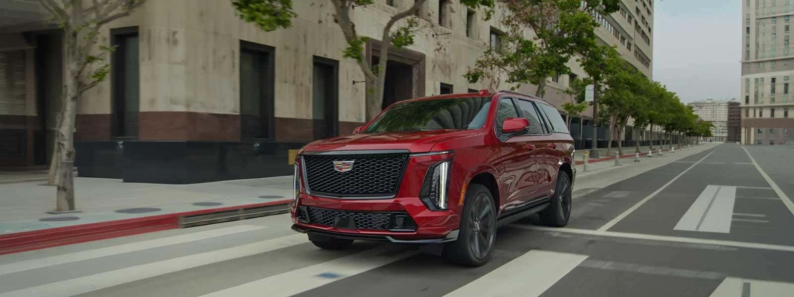 Red Cadillac Escalade driving on city street with buildings and trees in background, front grille and lighting clearly visible