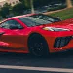 Red Corvette coupe driving on a city road with black wheels, side intake, and low front fascia.
