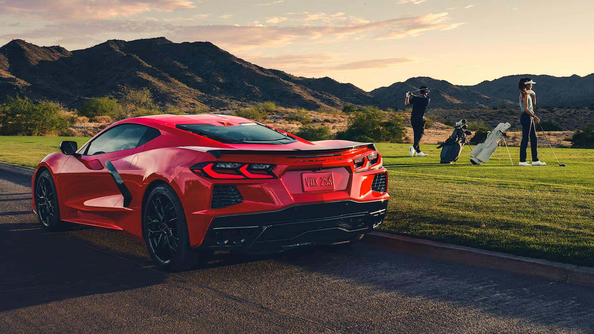 Red Corvette Stingray parked by a golf course with golfers, golf bags, grass, mountains, and a golden sky