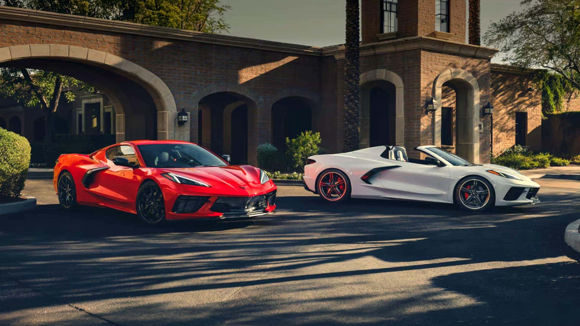 A red and a white Corvette Stingray parked outside a brick building with arched entryways