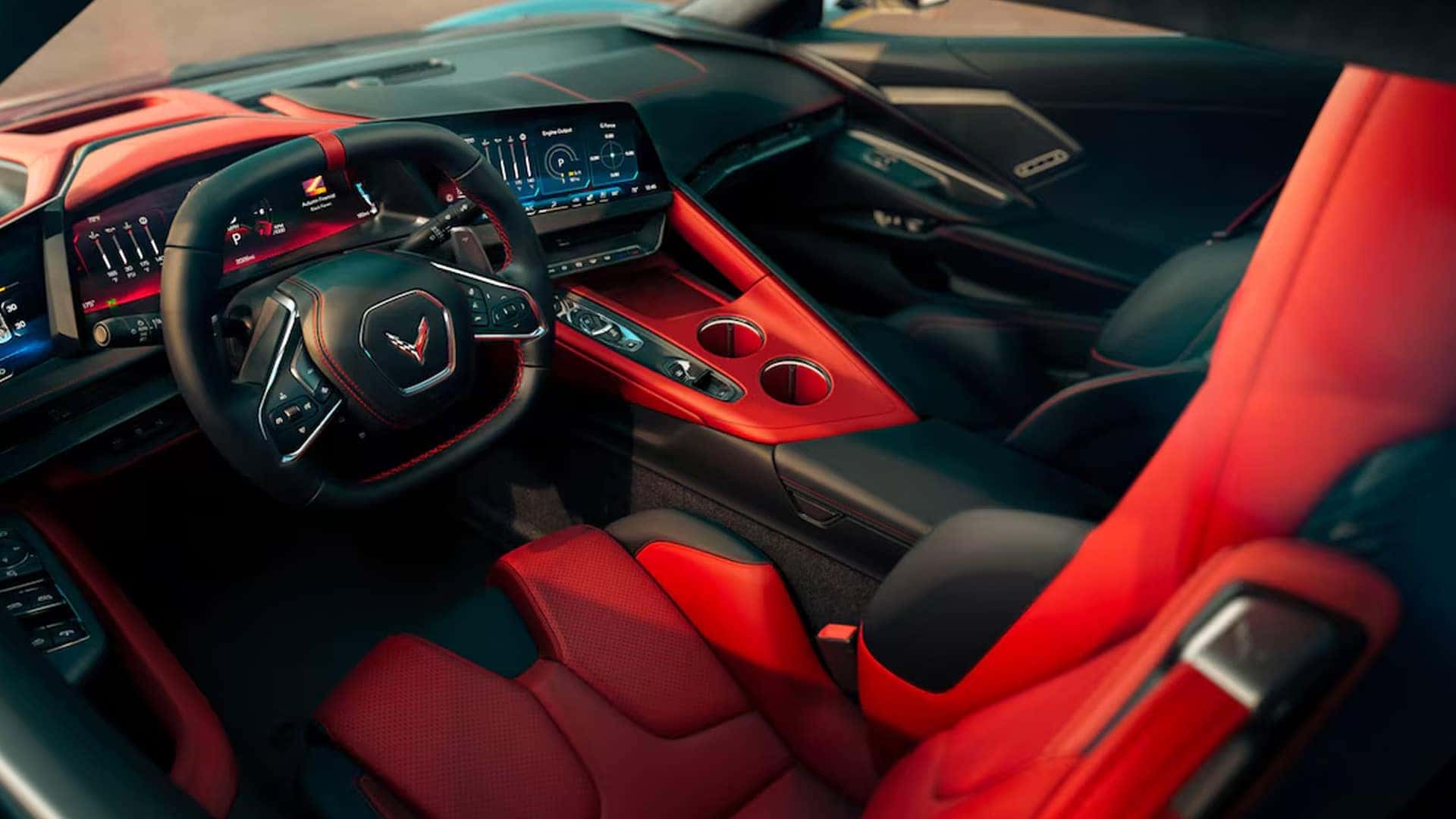 Angled view of Corvette cabin showing red and black sport seats, steering wheel, screens, and center console.