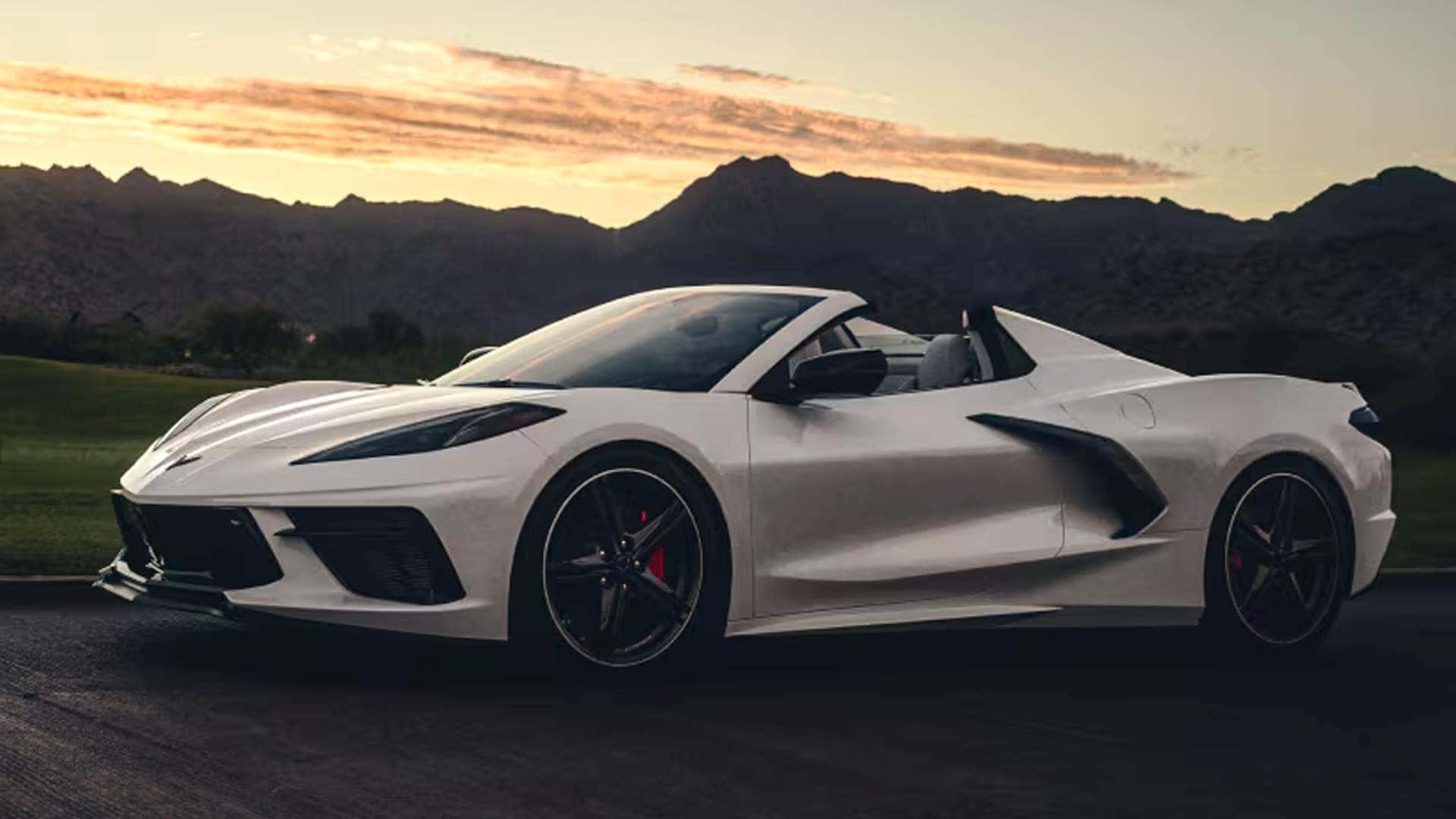 White Corvette convertible shown in profile with the roof open against a mountain sunset background.