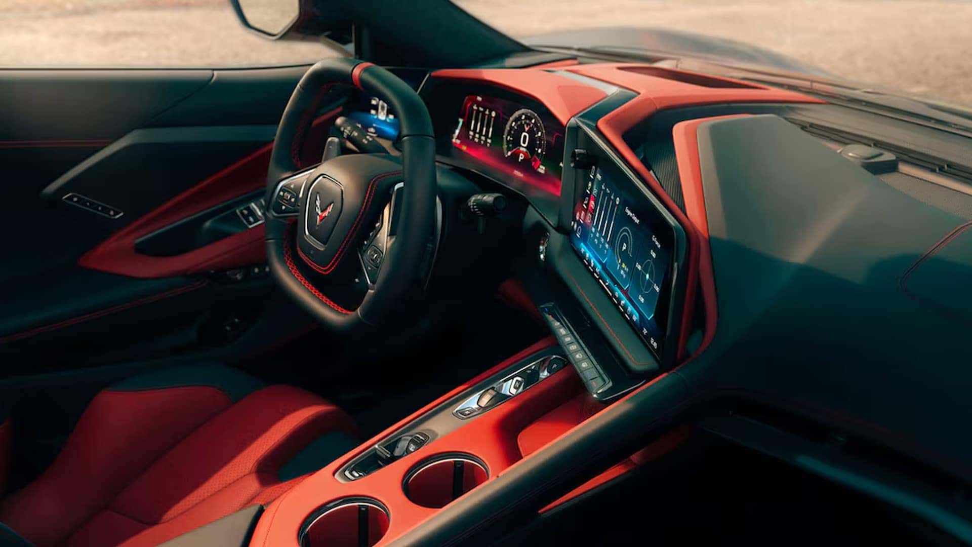 Wide view of Corvette cockpit with red and black trim, steering wheel, digital screens, console, and cupholders.