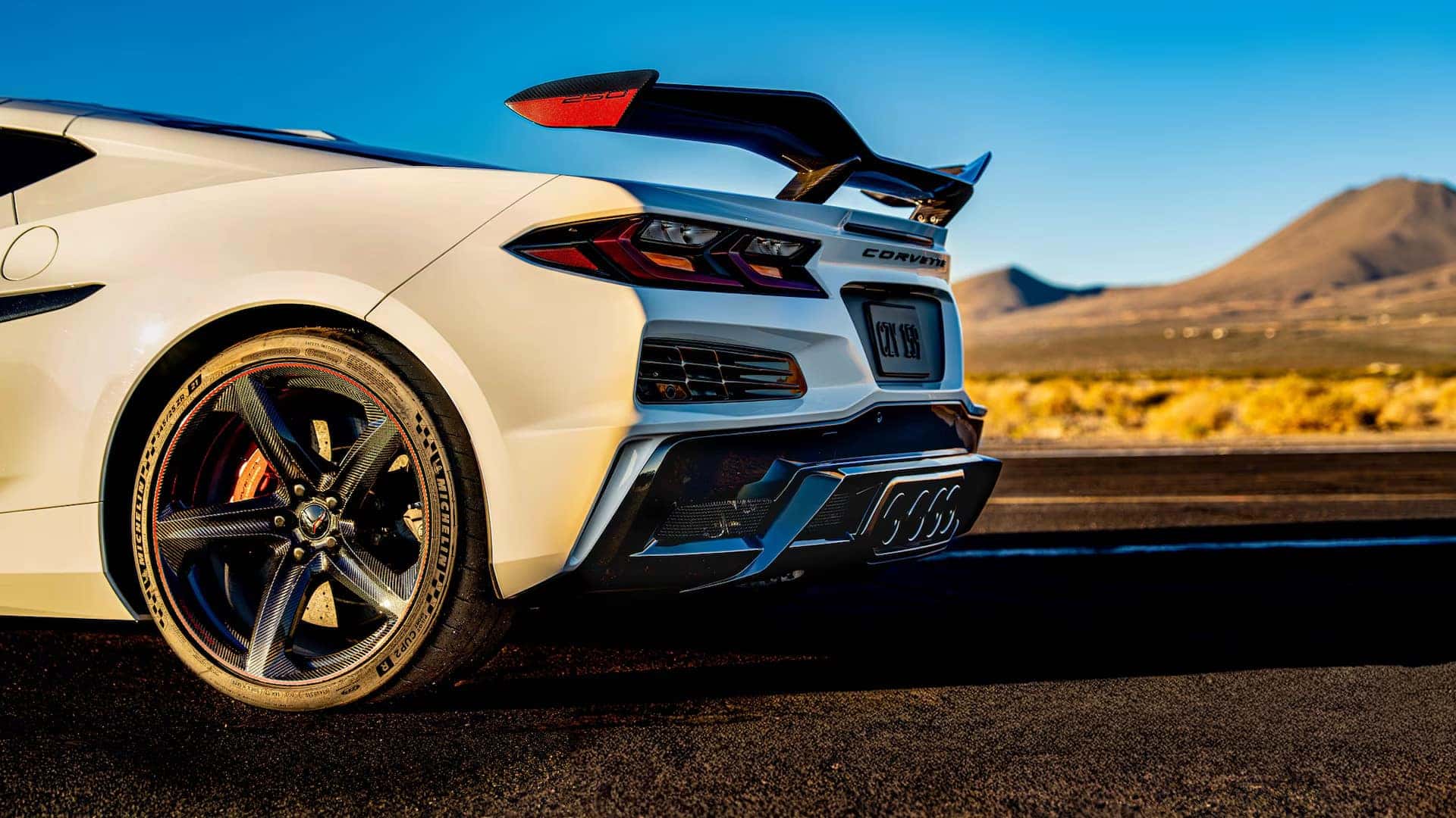 Rear close-up of a white Corvette with carbon fiber wheel, rear wing, taillight, and center exhaust.