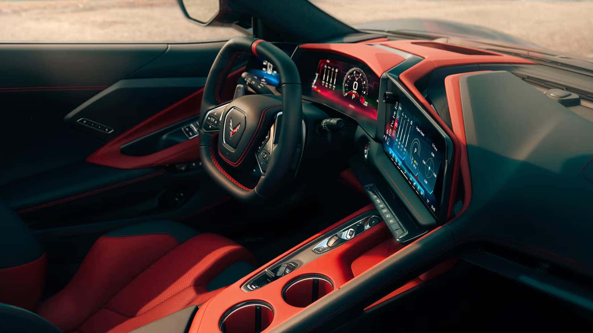 Red and black Corvette interior showing steering wheel, center console, touchscreen, and cupholders from driver view