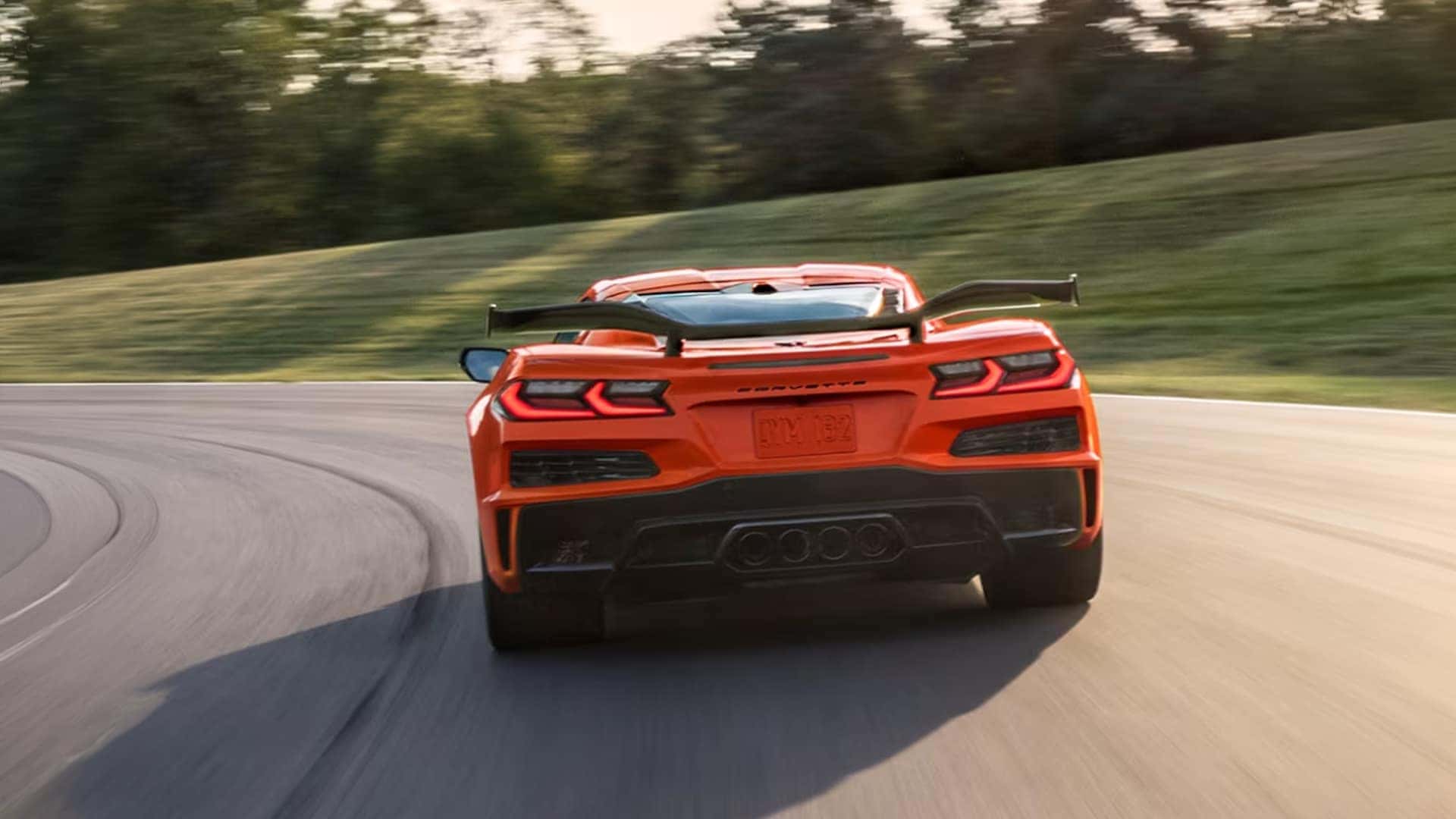 Rear view of orange Corvette Z06 driving on a curved road with visible taillights and large rear wing in motion