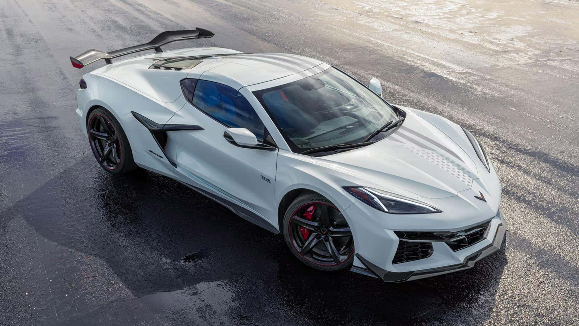White Corvette Z06 with rear wing parked on wet pavement, showing black wheels, red brake calipers, and hood stripe detail
