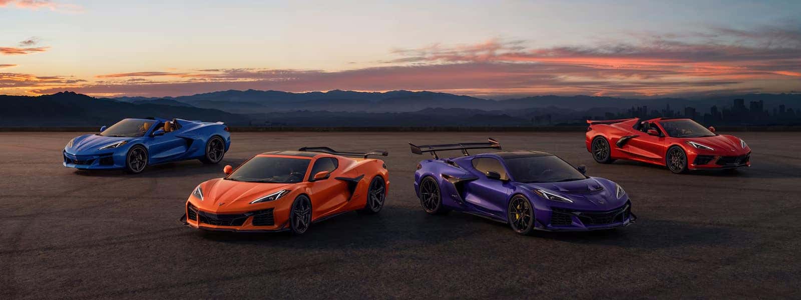Four Corvette models in different colors parked on open pavement at sunset with city skyline faintly visible in distance
