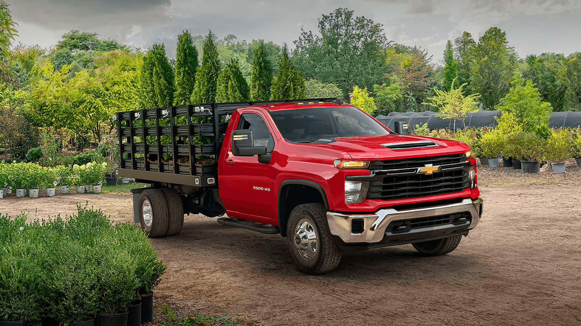Red Chevy Silverado 3500 HD flatbed truck parked on dirt lot surrounded by plants and trees in a nursery setting