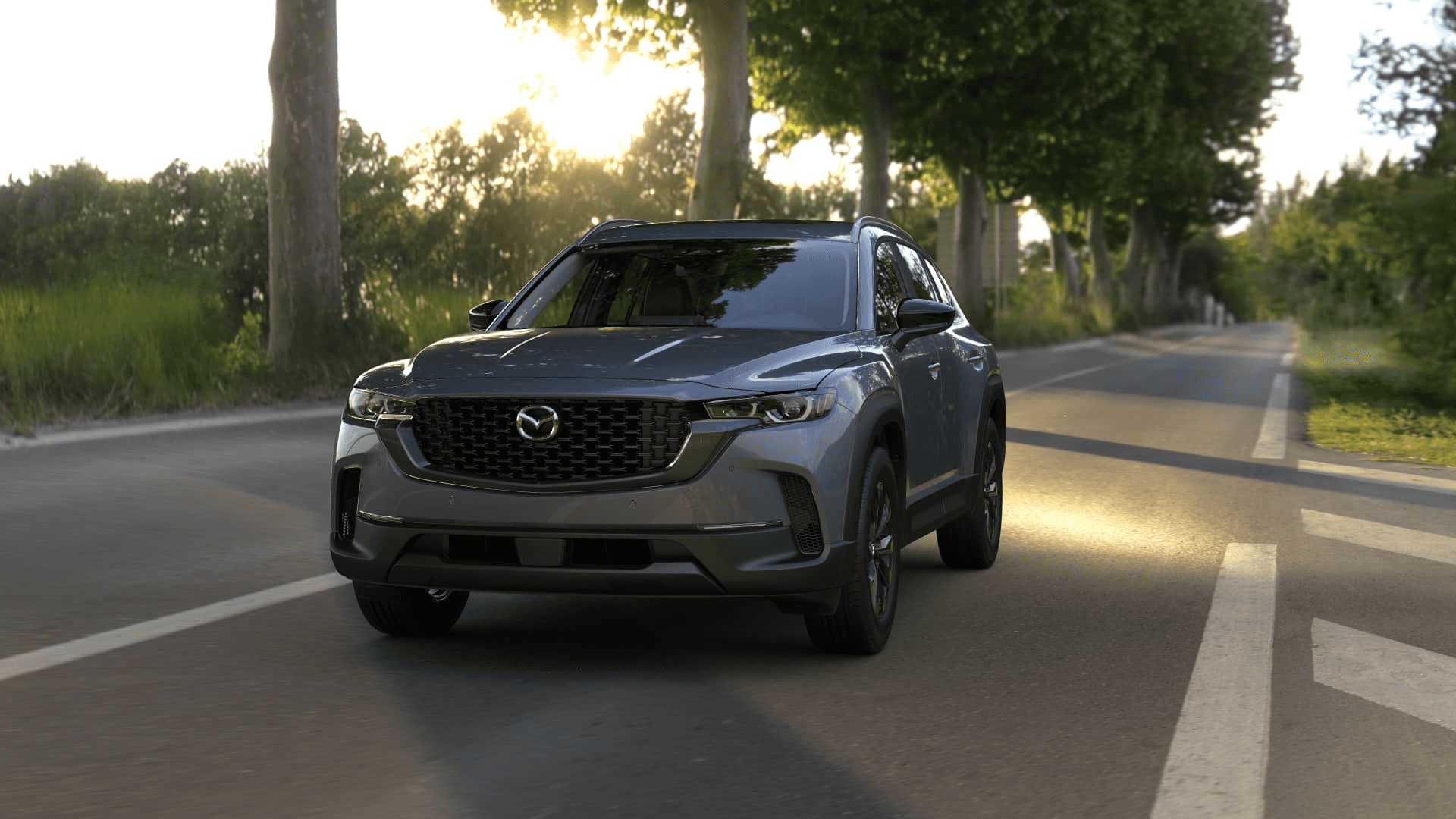 Gray Mazda CX-50 driving on a tree-lined road at sunset, shown from the front with grille and roof rails visible