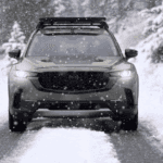 Front view of a Mazda CX-50 on a snowy road with headlights on, falling snow, and a roof rack above
