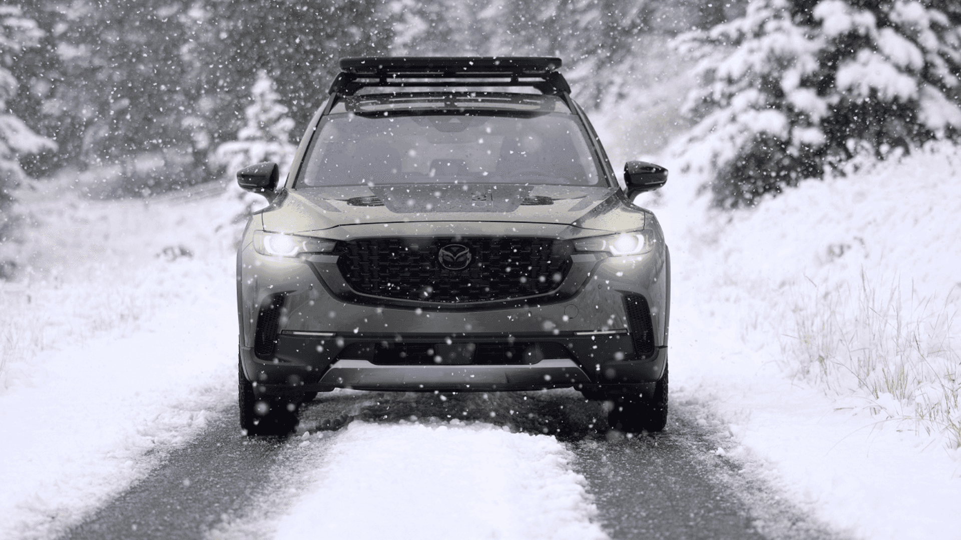 Front view of a Mazda CX-50 on a snowy road with headlights on, falling snow, and a roof rack above