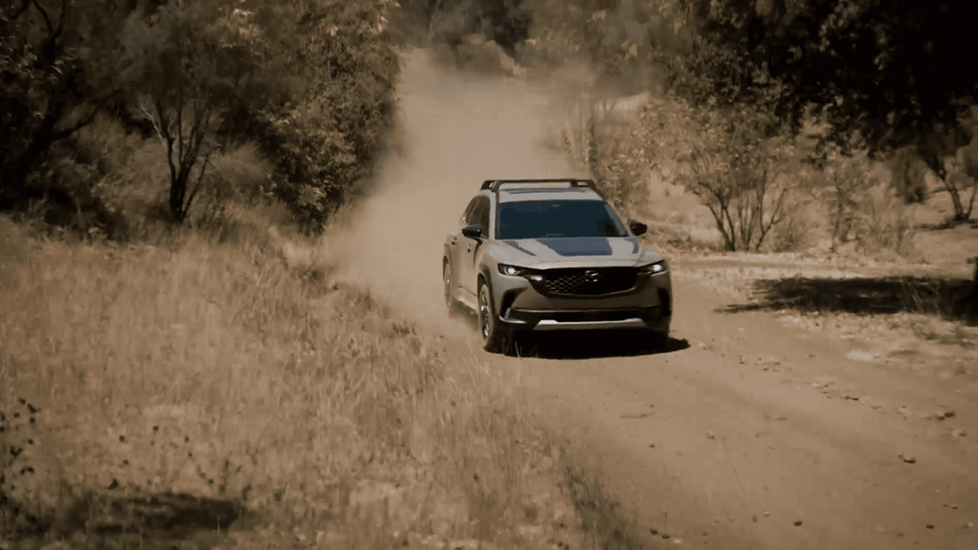 Mazda CX-50 driving on a dusty dirt road with trees and dry grass on both sides of the trail
