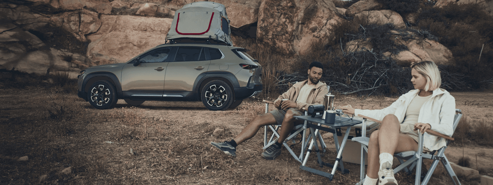 Mazda CX-50 parked near rocks with a rooftop tent, while two people sit at a camp table beside it