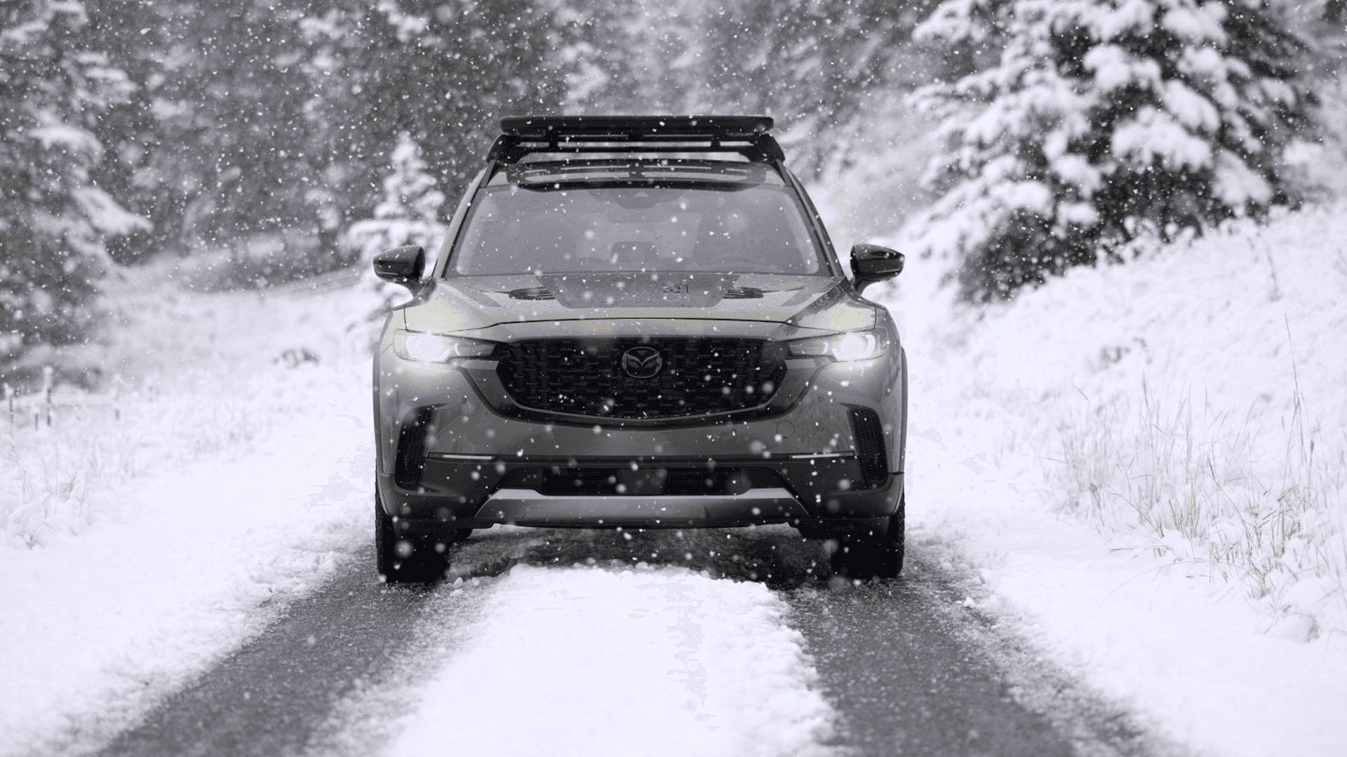 Front view of a gray Mazda CX-50 driving on a snow-covered road with falling snow and trees on both sides