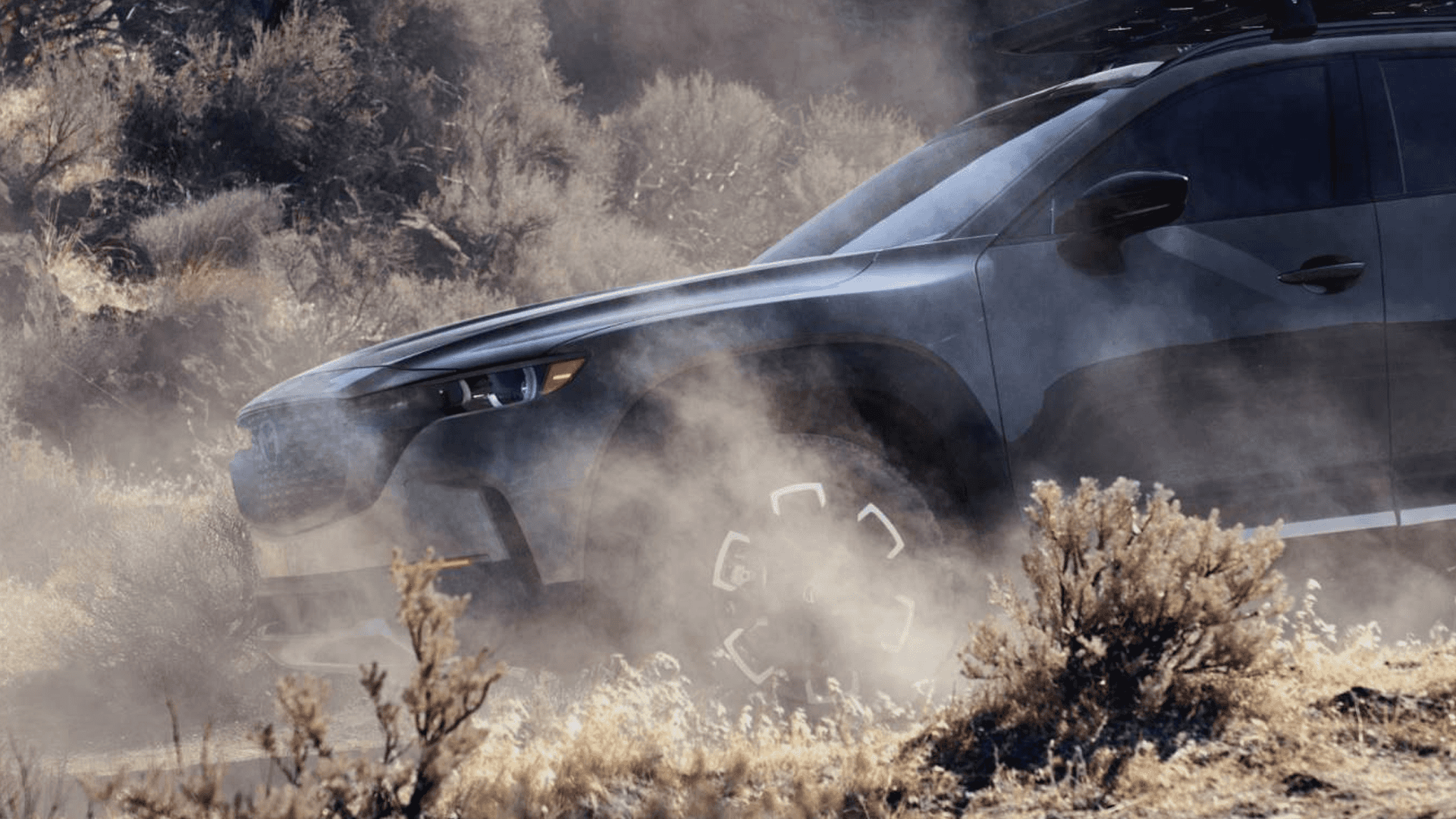 Dark Mazda CX-50 driving through a dusty off-road landscape with brush and sunlight around the front side