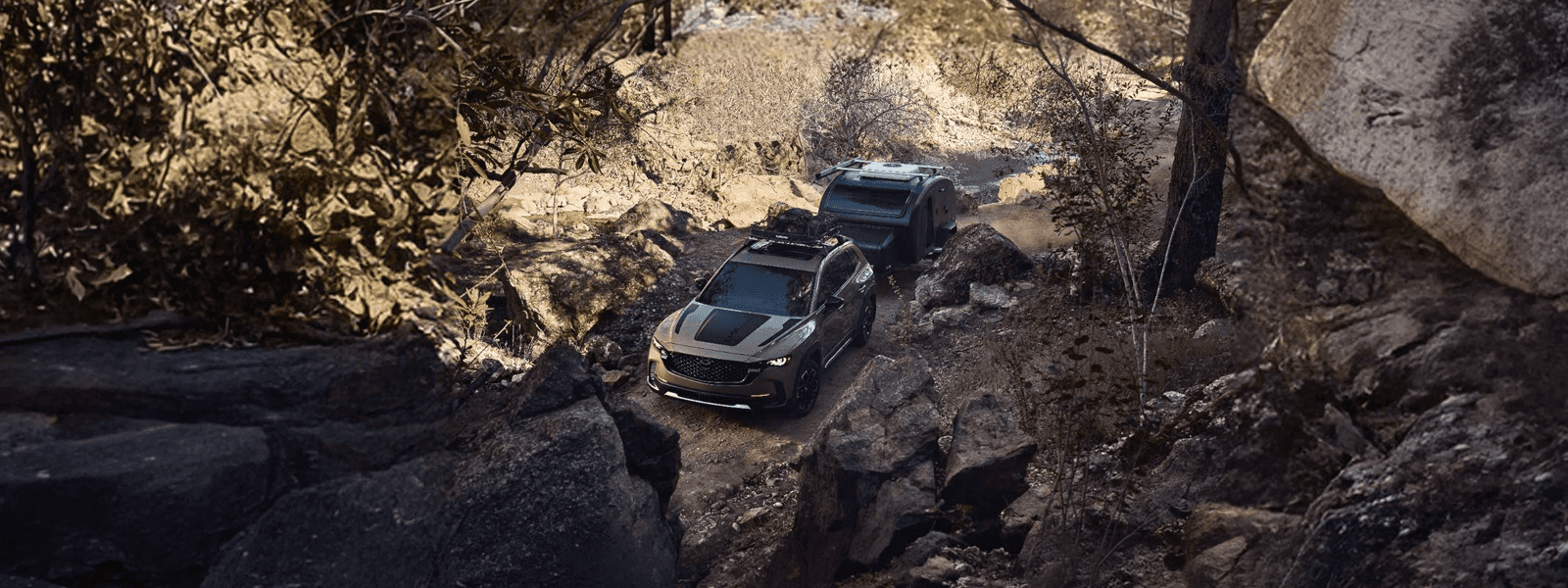 Mazda CX-50 towing a small trailer on a rocky forest trail with roof gear and large boulders surrounding it