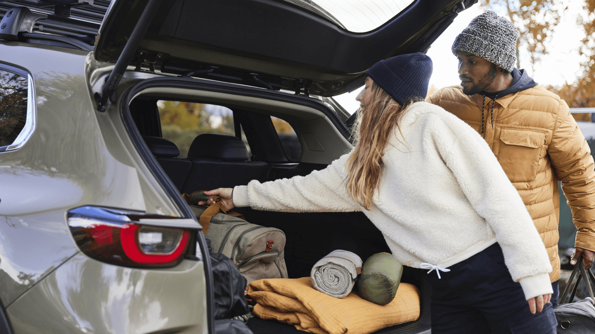 Two people loading bags and rolled blankets into the open rear cargo area of a light-colored SUV