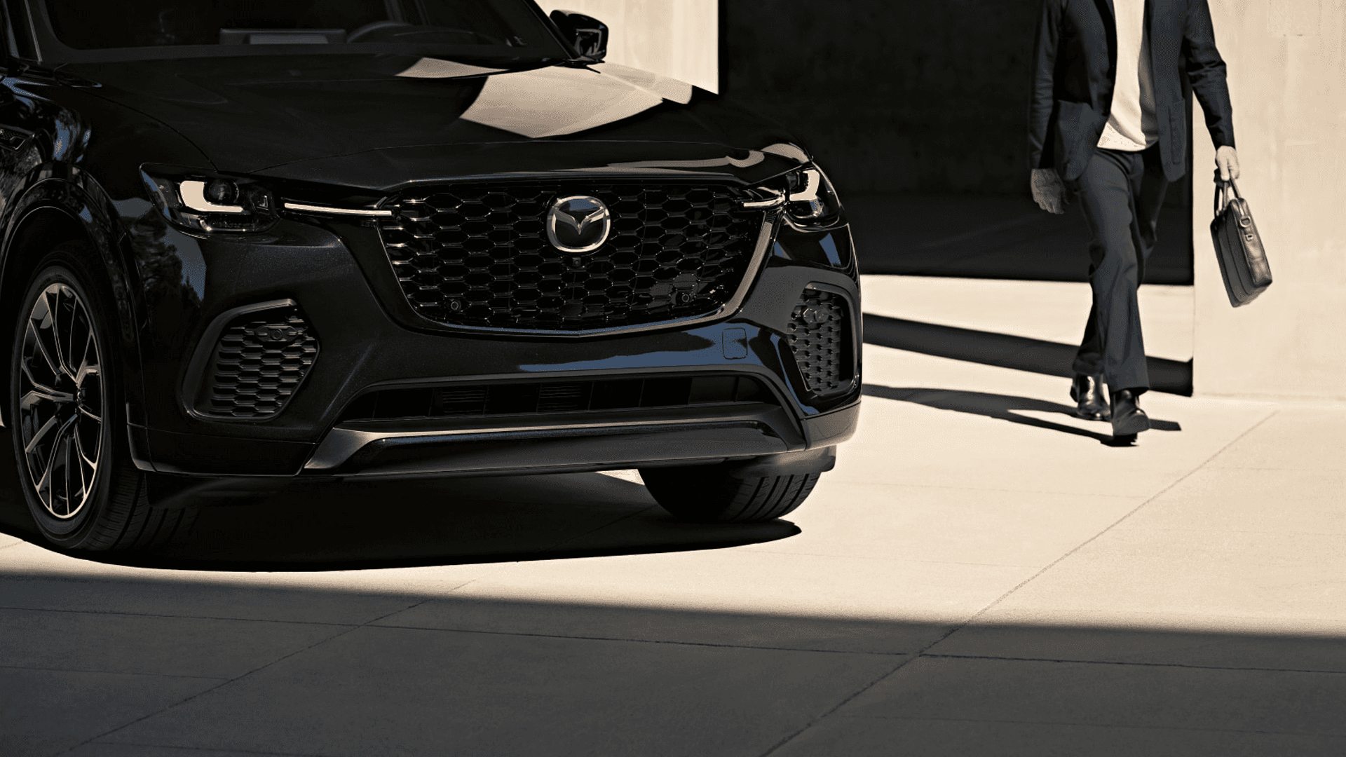 Front view of a black Mazda SUV with grille and headlights visible, while a person walks nearby holding a briefcase