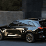 Black Mazda SUV driving on a city street with reflections on body panels and blurred pedestrian in background