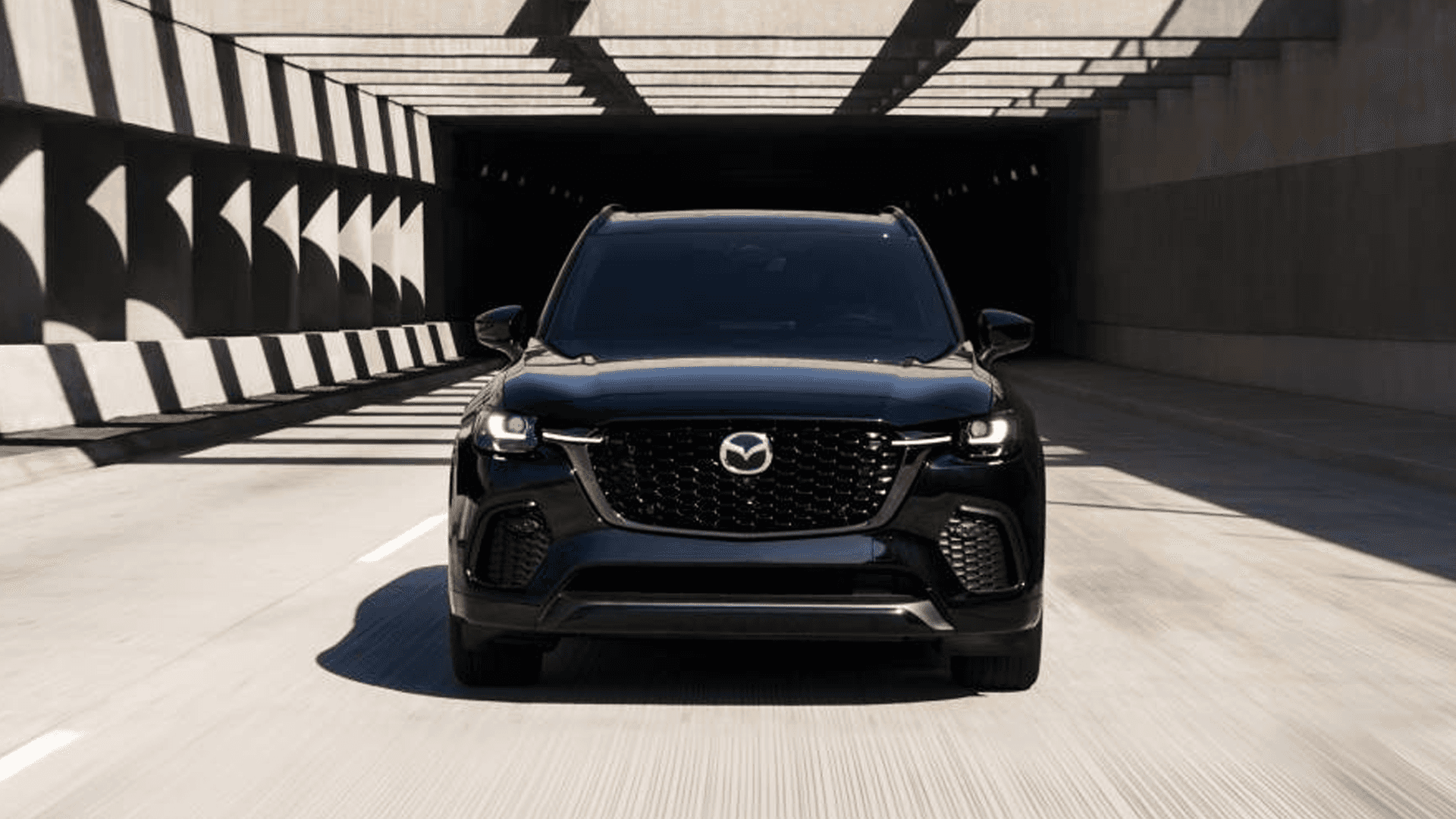 Front view of a black Mazda CX-70 driving through a tunnel with strong shadow patterns on the walls