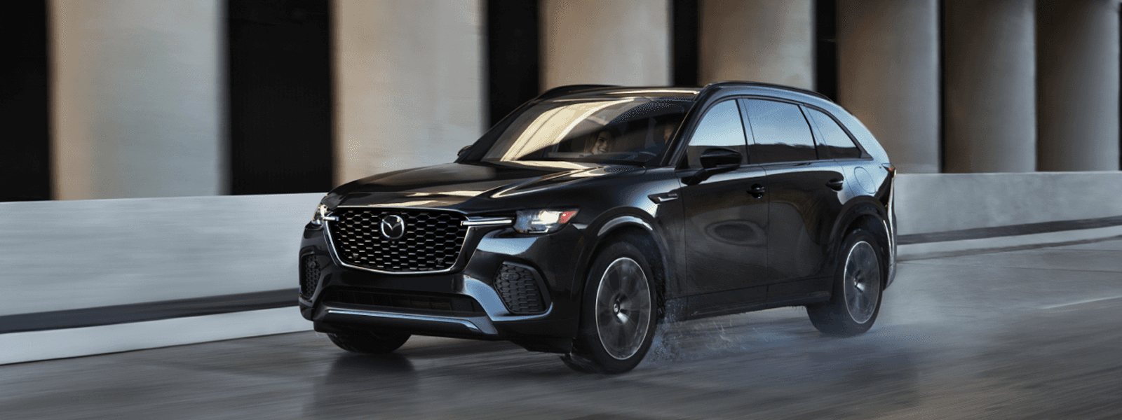 Black Mazda CX-70 driving on a wet roadway beside large concrete columns and a curved barrier