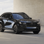Three-quarter view of a black Mazda CX-70 parked in an open concrete area with a modern building in the background