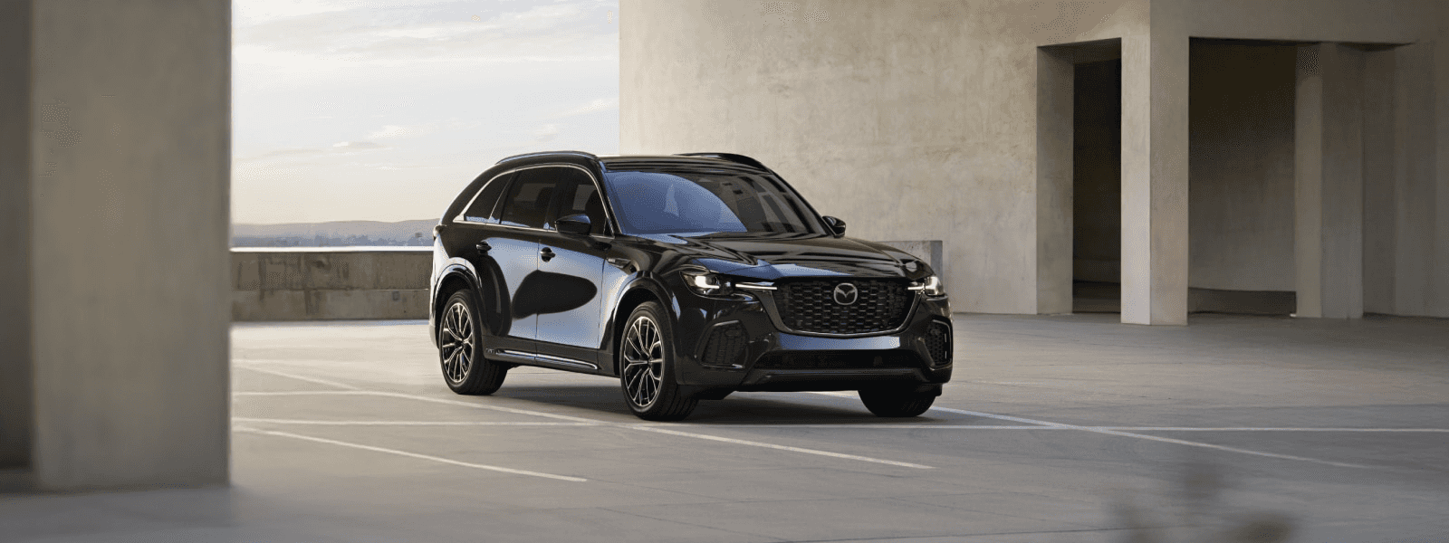 Three-quarter view of a black Mazda CX-70 parked in an open concrete area with a modern building in the background